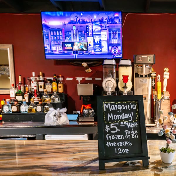A bar area with a "Margarita Monday" sign, liquor bottles, beer taps, and a TV showing a city scene.
