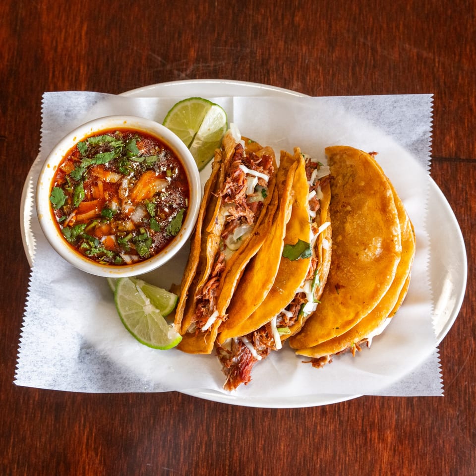 Three birria tacos with shredded meat, cheese, and cilantro, served with a side of consommé and lime wedges.