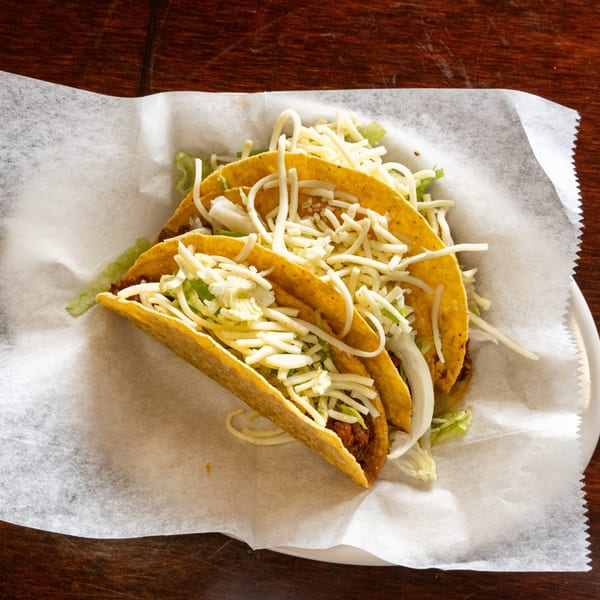 Three crispy tacos filled with meat, shredded lettuce, and cheese, served on a white paper-lined plate.