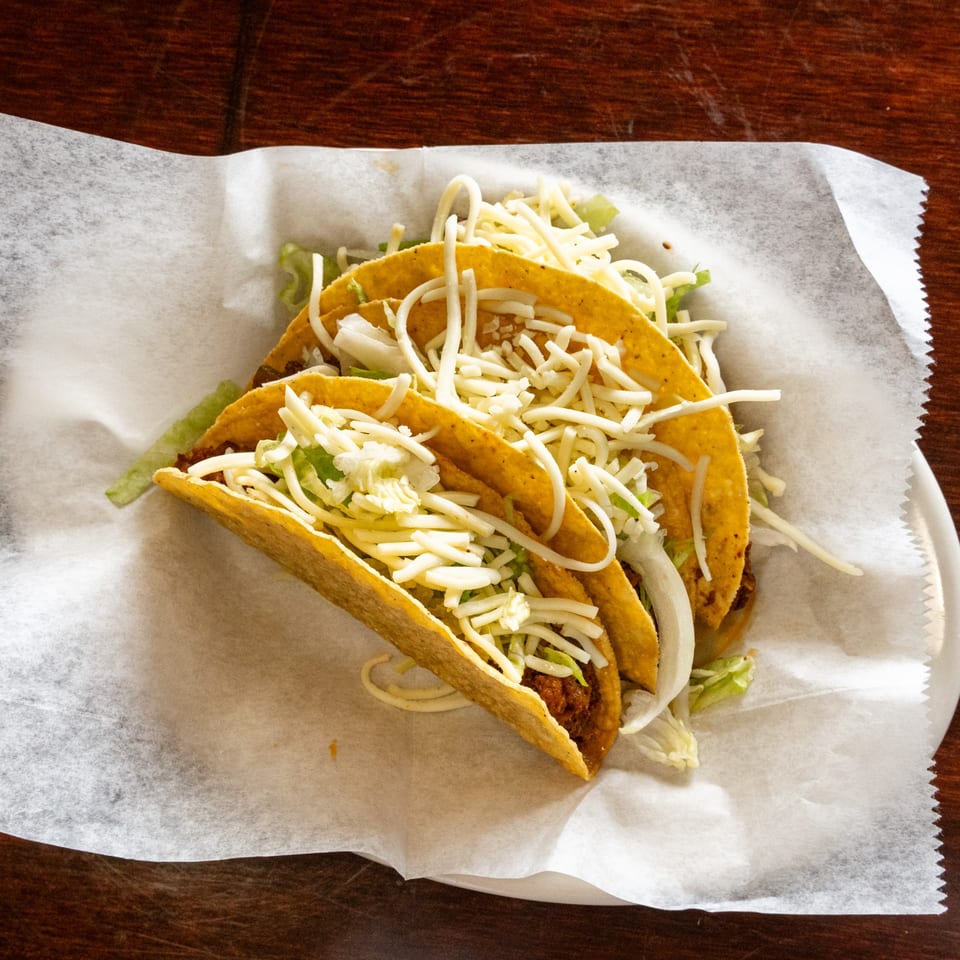Three crispy tacos filled with meat, shredded lettuce, and cheese, served on a white paper-lined plate.
