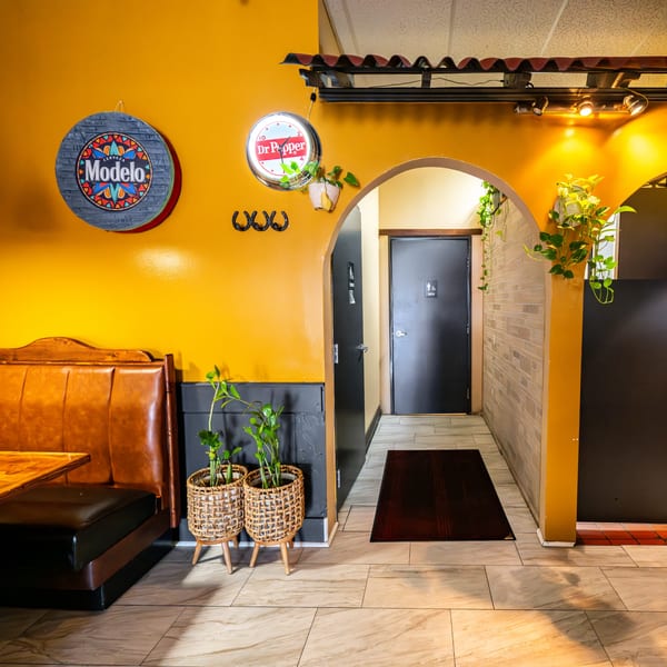 A vibrant yellow wall with a Modelo sign, Dr. Pepper clock, horseshoe decor, and an arched doorway to restrooms.