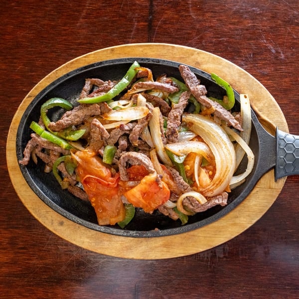 Sizzling beef fajitas with green peppers, onions, and tomatoes in a black skillet on a wooden board.