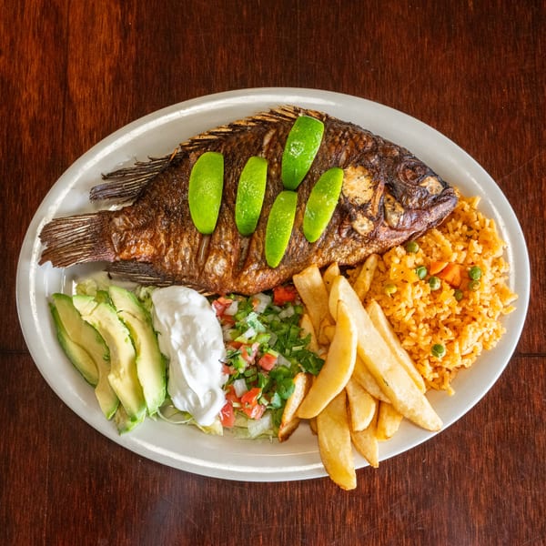 A whole fried fish with lime slices, rice, french fries, avocado, sour cream, and pico de gallo.