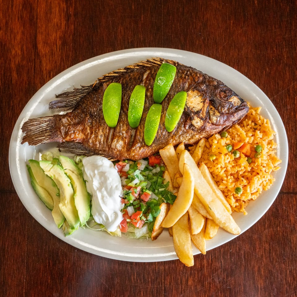 A whole fried fish with lime slices, rice, french fries, avocado, sour cream, and pico de gallo.