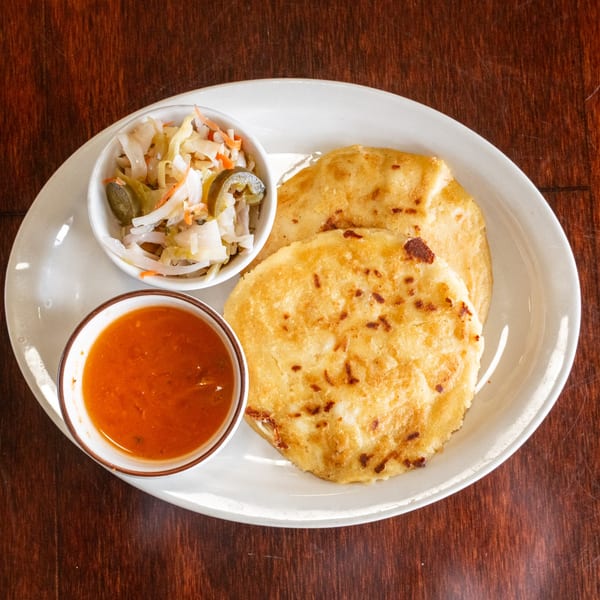 Two golden pupusas with a side of curtido and a bowl of red salsa on a white plate.