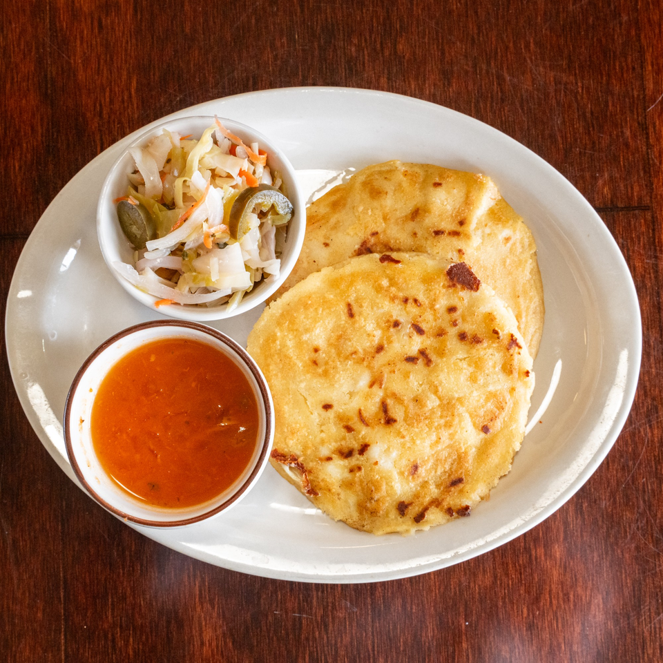 Two golden pupusas with a side of curtido and a bowl of red salsa on a white plate.