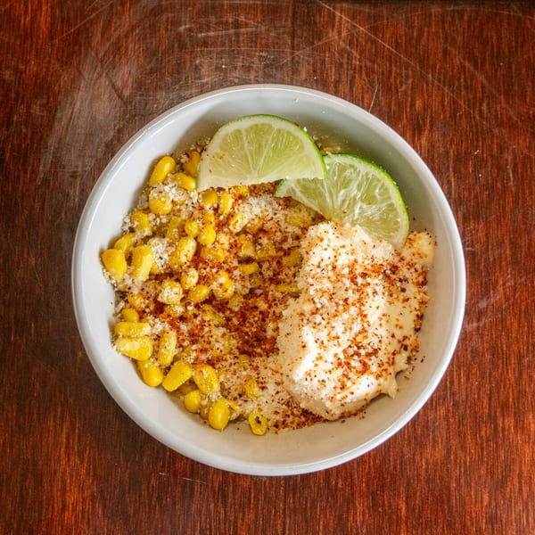 Elote in a white bowl, topped with cotija cheese, chili powder, and lime wedges.