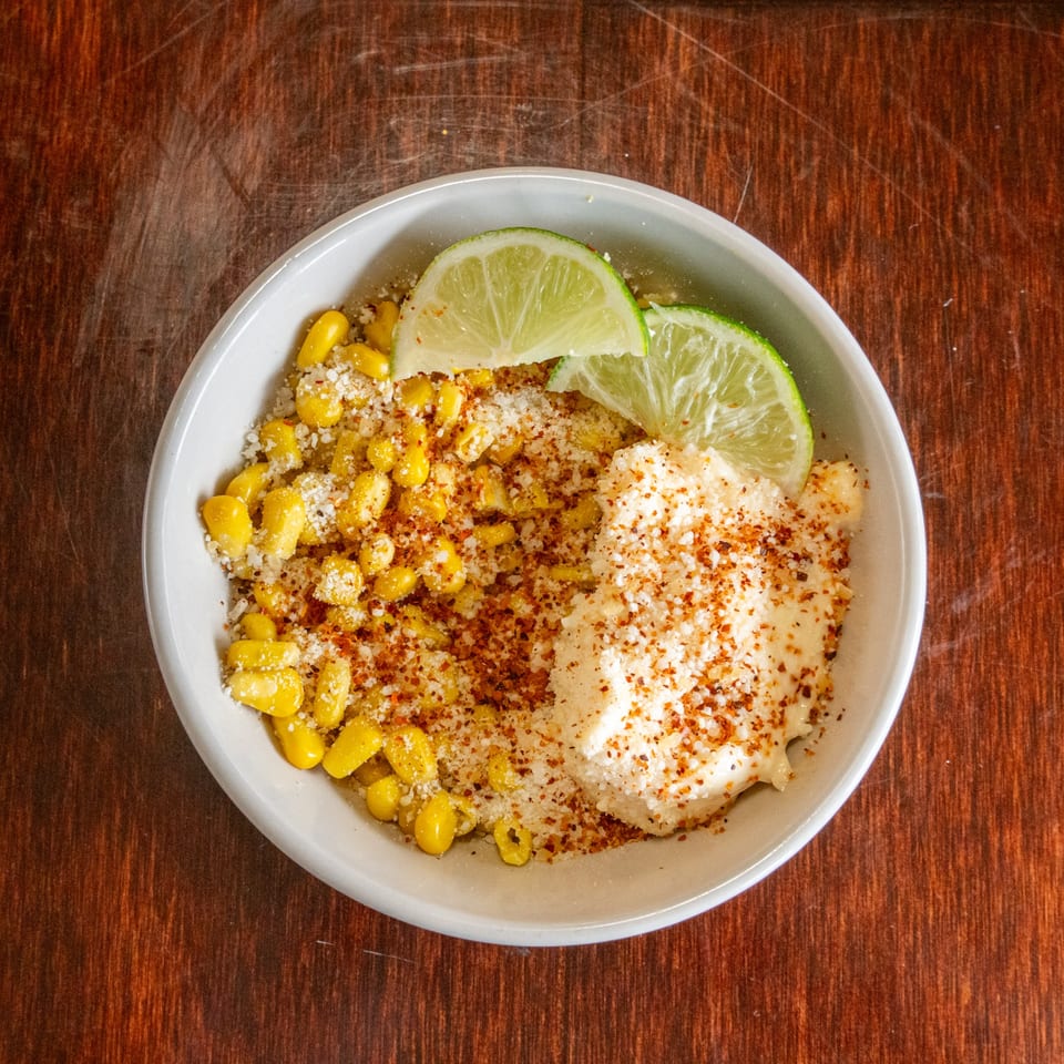 Elote in a white bowl, topped with cotija cheese, chili powder, and lime wedges.