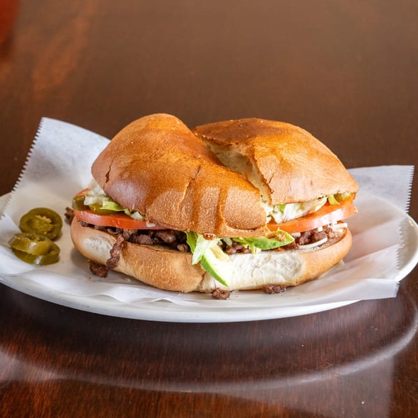 A Mexican torta sandwich with meat, lettuce, tomato, avocado, and onions, served with jalapeños.