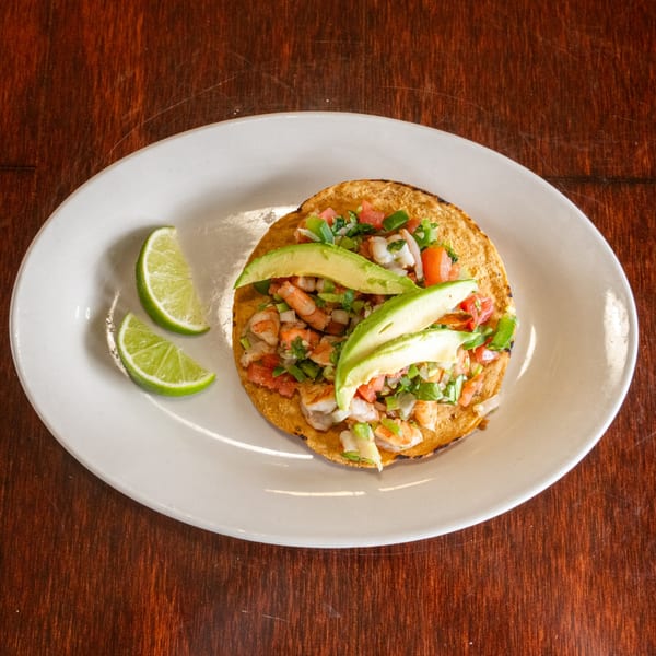 Shrimp tostada with diced tomatoes, onions, cilantro, and avocado slices, served with lime wedges.