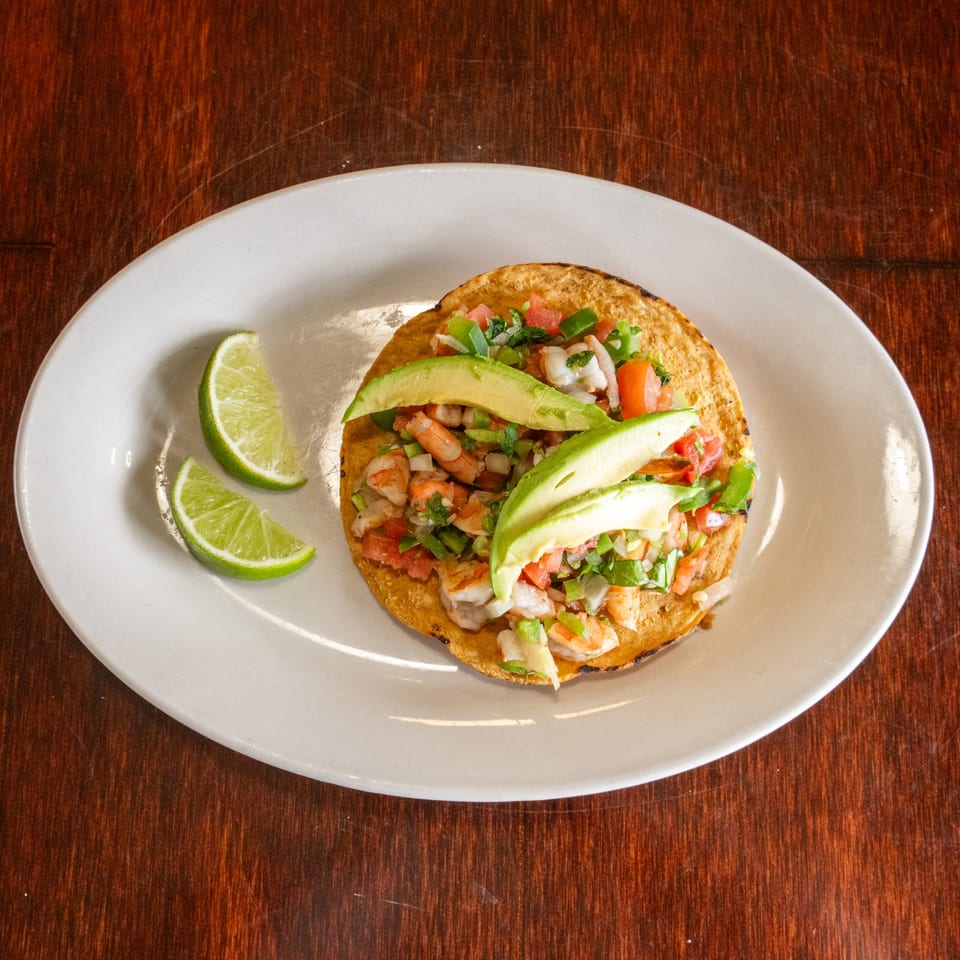 Shrimp tostada with diced tomatoes, onions, cilantro, and avocado slices, served with lime wedges.